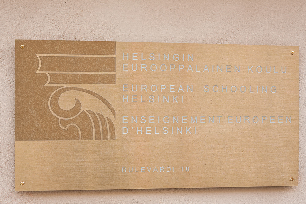 Kaiverrettu messinkikilpi, koulun nimikyltti. Tekstit kaiverrettu, kuvio hiekkapuhallettu. Viimeistelynä lakkaus.